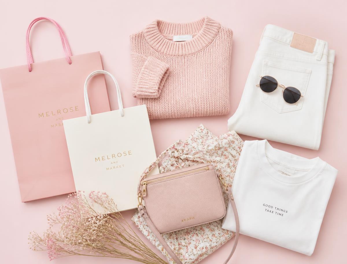 Pastel pink shopping bags, a folded sweater, jeans and a small handbag arranged on a blush background